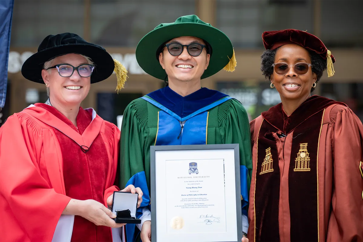 A graduate in green convocation gown holding his degree while 2 professors in red and brown gown stand on either side of him