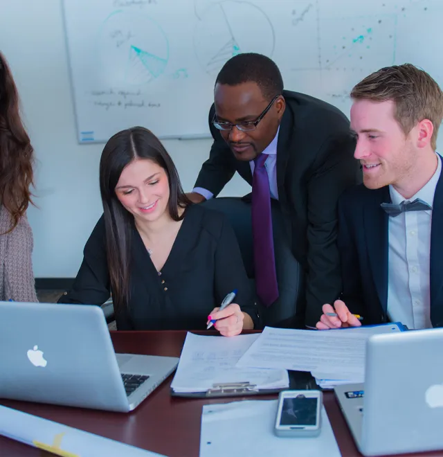 A group of students and a professor in business attire looking at a laptop in a work setting