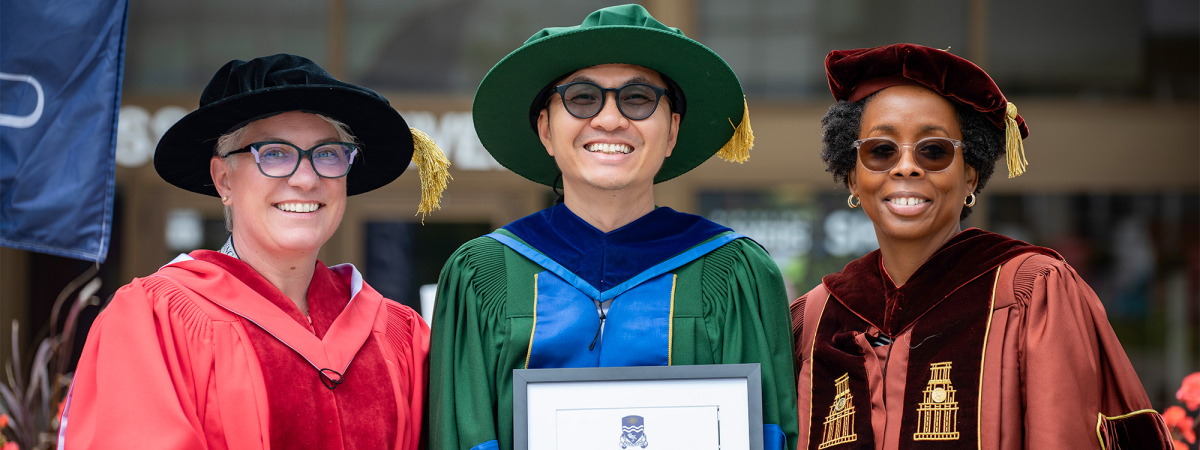 A graduate in green convocation gown holding their degree with 2 professors on both sides of him