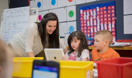 A student teacher helping 2 students with something on the ipad in a classroom setting