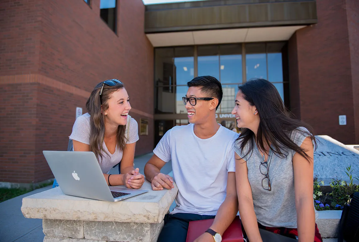 Three students sit on bench outside the front entrance to Nipissing University.