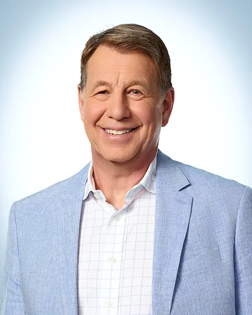 A man in light blue blazer against a light background with blue corner vignette